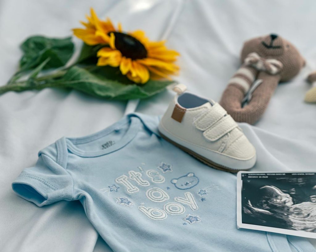 A charming baby announcement featuring a onesie, ultrasound, shoe, and sunflower on a soft blue background.
