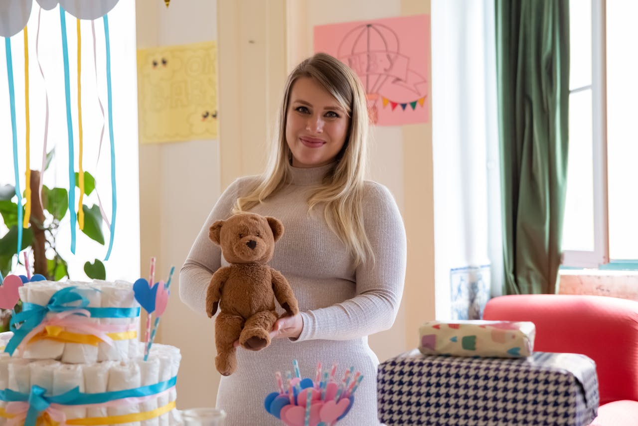 Smiling pregnant woman at a baby shower holding a teddy bear in a decorated room.
